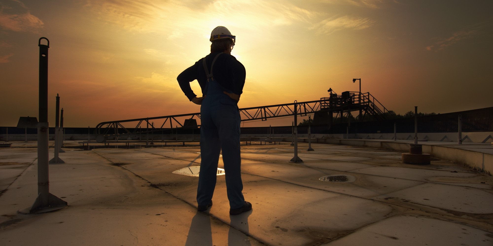 Man In Safety Clothing Standing On Top Of Oil Tank At Sunrise Rear View